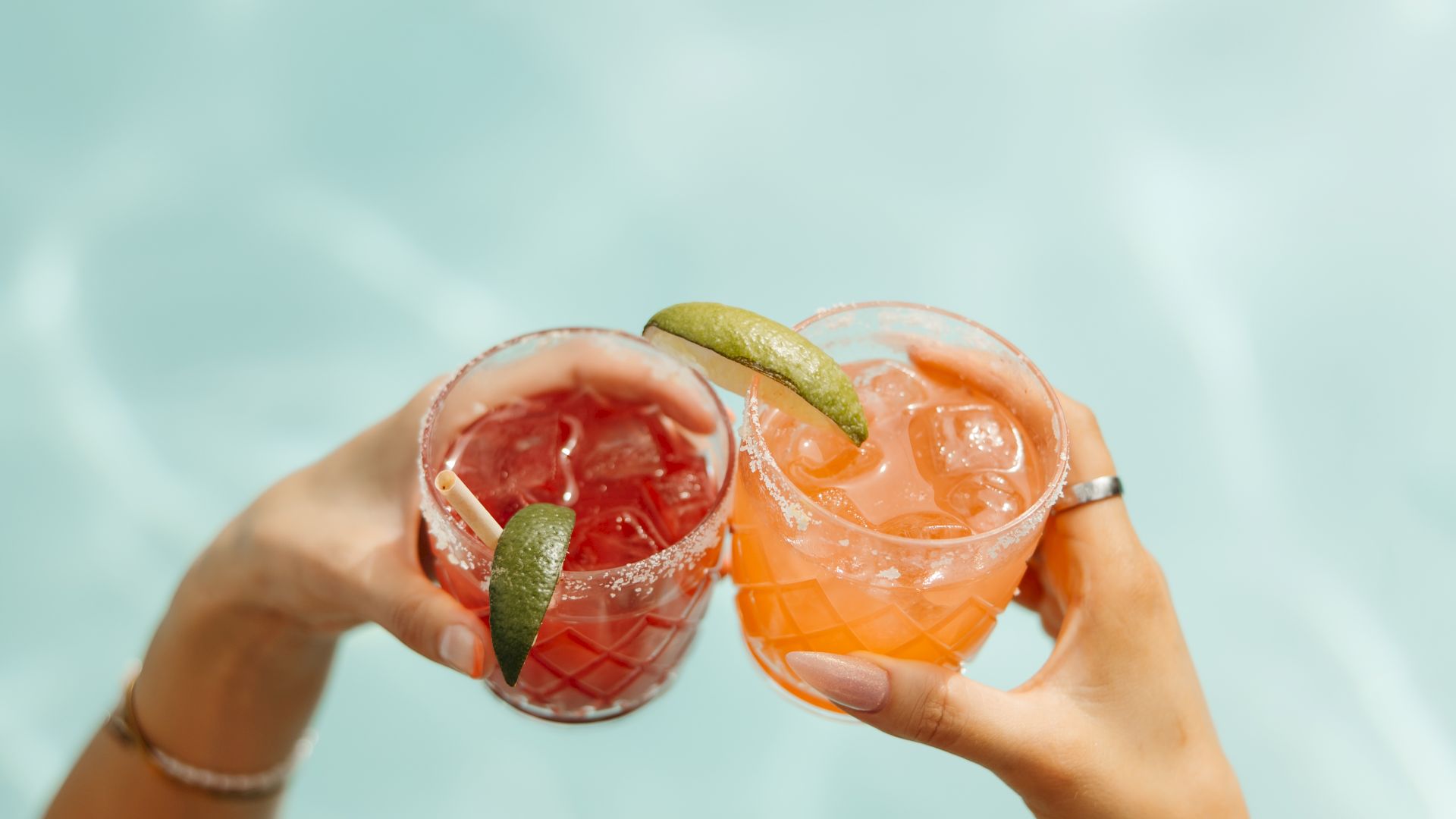 Two hands hold up glasses with colorful drinks garnished with lime slices, set against a backdrop of water, possibly a pool.