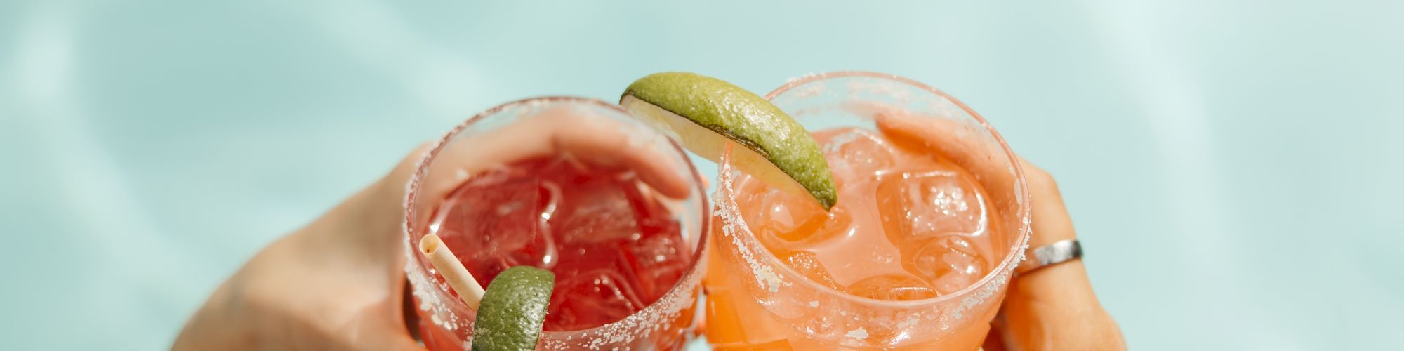 Two hands hold up glasses with colorful drinks garnished with lime slices, set against a backdrop of water, possibly a pool.