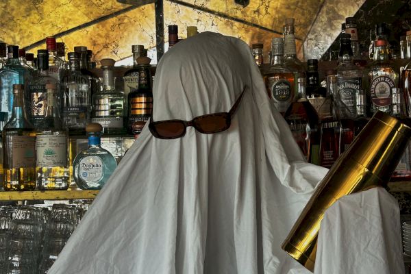 A person dressed as a ghost with sunglasses holds a cocktail shaker in front of a bar stocked with various bottles.