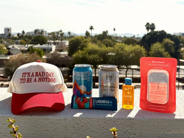 A collection of items on a ledge: a red-and-white cap reading &ldquo;IT'S A BAD DAY TO BE A HOT DOG,&rdquo; cans of beer, a small bottle of sunscreen, a blue flask, a pouch labeled &ldquo;LIQUID LITE,&rdquo; and a red emergency kit on a sunny rooftop with trees in the background.