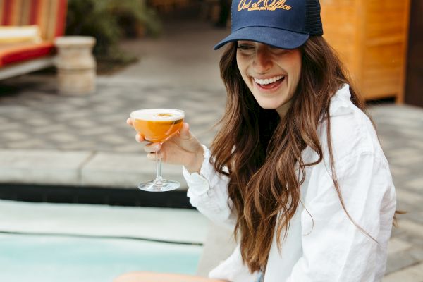 A woman in a white shirt and cap sits by a pool, smiling and holding a drink. Another drink is on the ground nearby.