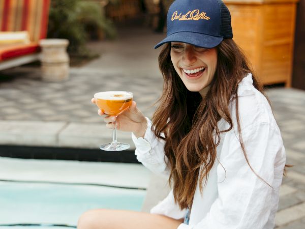 A woman in a white shirt and cap sits by a pool, smiling and holding a drink. Another drink is on the ground nearby.