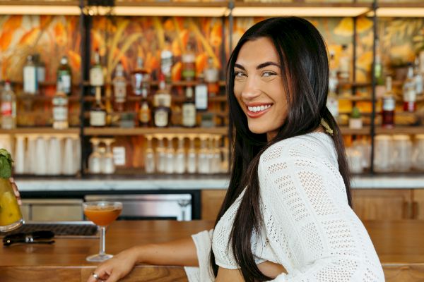 A woman in a white outfit sits at a bar, holding a cocktail, with a smile, and a tropical decor background.