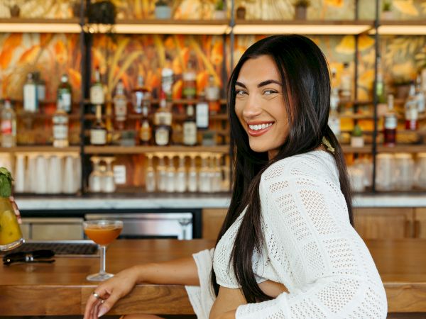 A woman in a white outfit sits at a bar, holding a cocktail, with a smile, and a tropical decor background.