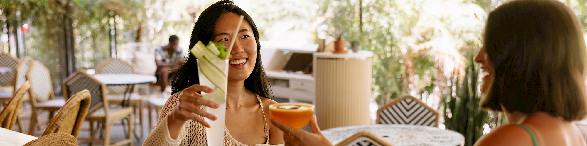 Two people are sitting at a bar, clinking glasses in a cheerful setting with outdoor furniture and greenery around them.