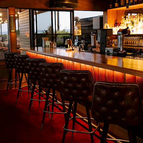 A cozy bar with a wooden counter, warm lighting, and tufted leather stools lined up along the bar, bottles and glassware in the back.