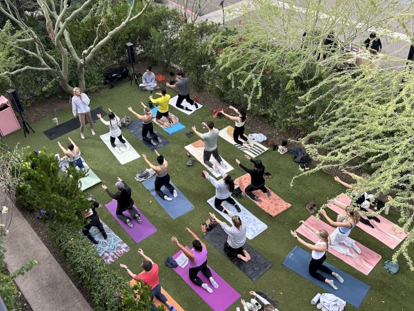 Yoga class taking place on colorful mats in a courtyard garden, participants strike poses on mats amid green trees and urban surroundings.