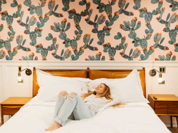 Decorative cactus-pattern wallpaper above a bed; a woman lounges on the white bedding between two wooden nightstands in a cozy hotel room.