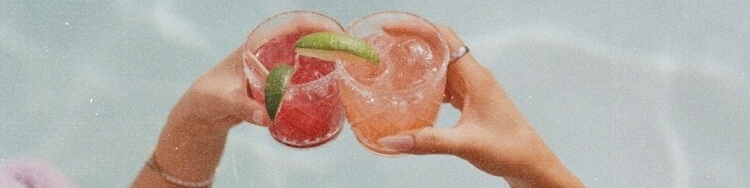 Two people share a pink grapefruit-like fruit by a pool, toasting it across the water as sunlight ripples in the background.