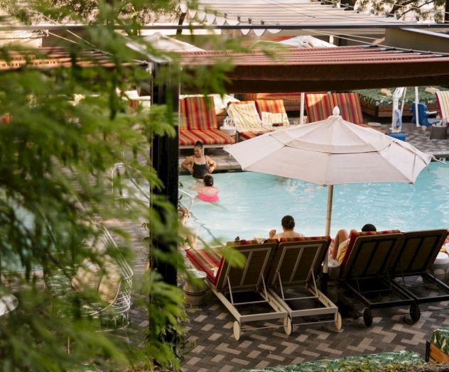 Poolside lounge area with chairs, umbrellas, and a turquoise pool; trees frame the scene as guests relax in a sunny resort setting.