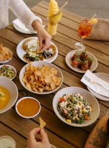 A table full of chips and bowls of colorful dishes, with hands grabbing nachos, a creamy dip, corn salsa, and drinks in a sunny outdoor setting.