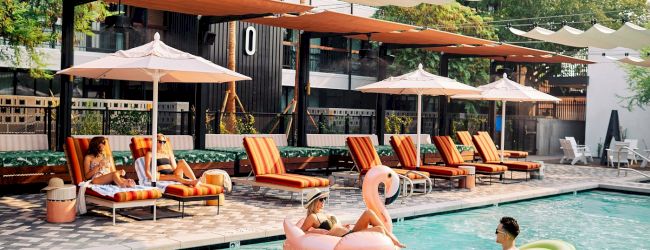 A sunny hotel pool scene with orange loungers, umbrellas, and people floating on a pink flamingo and a green inner tube in the water.