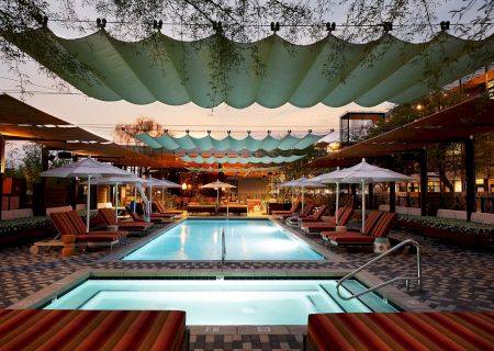 A stylish poolside retreat with a rectangular main pool, shaded lounge beds, cabanas, and an inviting turquoise spa, under decorative canopies.