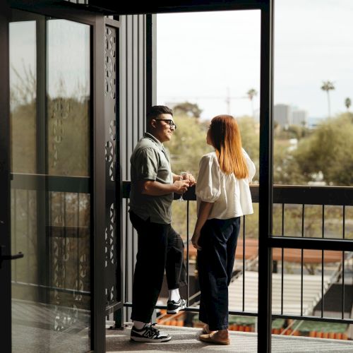Two people stand on a balcony, chatting and facing each other as the door opens to an outdoor view with trees and buildings in the distance.
