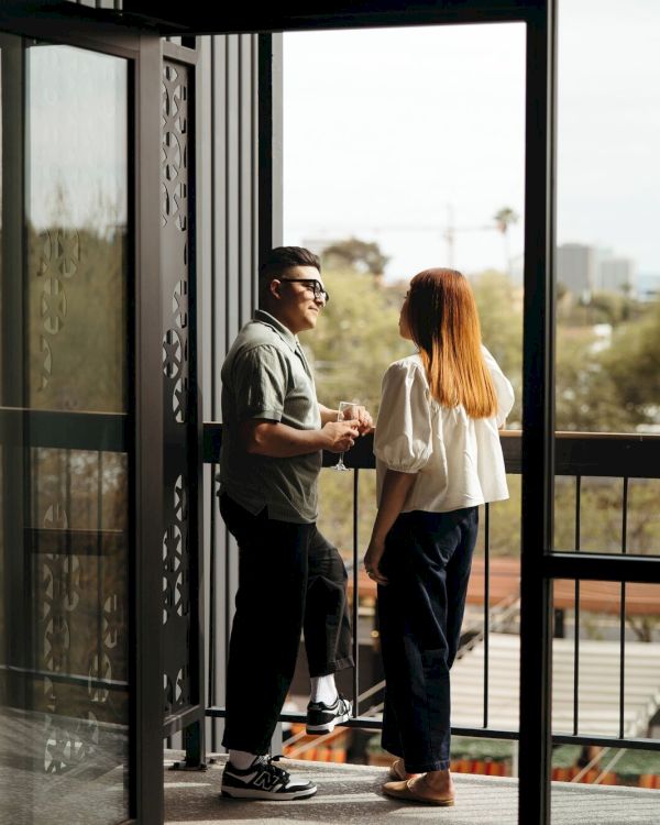 Two people stand on a balcony, chatting and facing each other as the door opens to an outdoor view with trees and buildings in the distance.