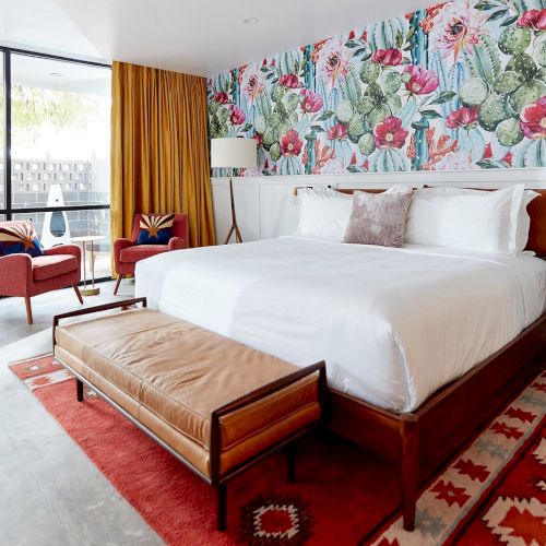 A bright hotel room with a tropical floral accent wall above the bed, white bedding, brown wood furniture, a red rug, and a tan bench at the foot of the bed. The space opens to a living area with red chairs and large glass doors.