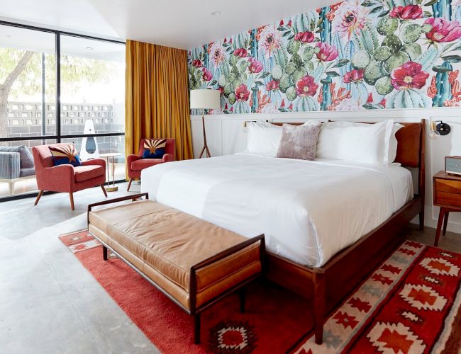 A bright hotel room with a tropical floral accent wall above the bed, white bedding, brown wood furniture, a red rug, and a tan bench at the foot of the bed. The space opens to a living area with red chairs and large glass doors.
