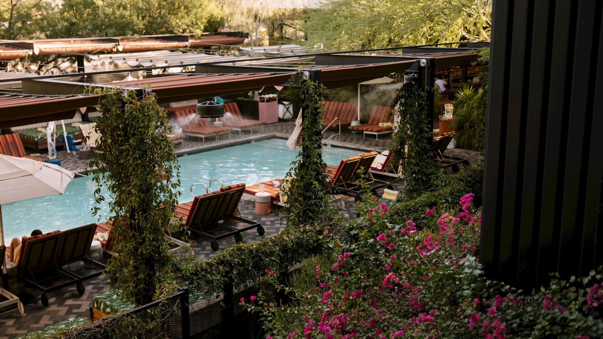 An outdoor resort patio with a heated pool, red-striped loungers, leafy vines and flowers, and a misty steamer under brown cabanas.