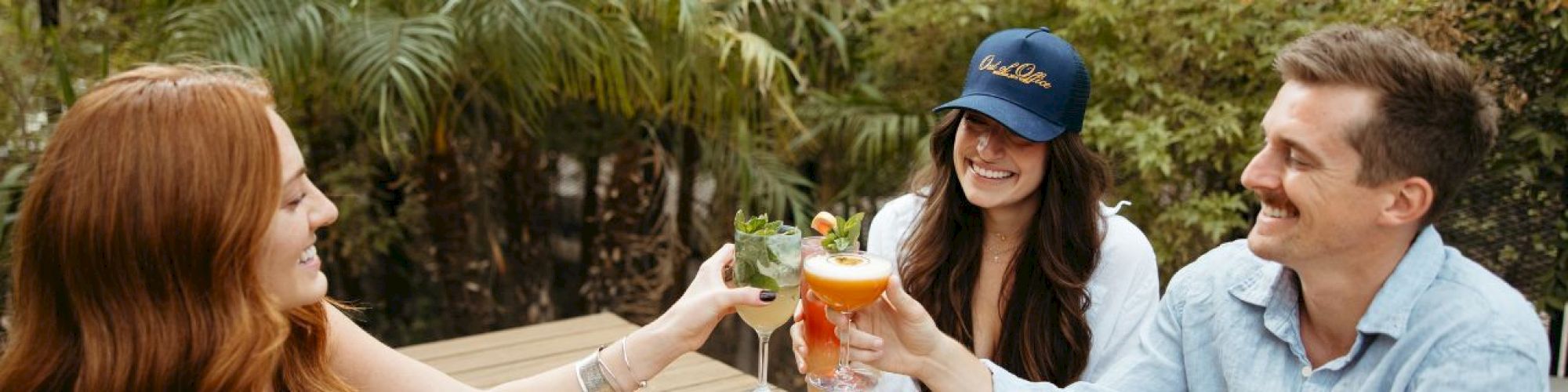 Three people sitting outside clinking glasses, enjoying drinks around a wooden table with lush greenery in the background.