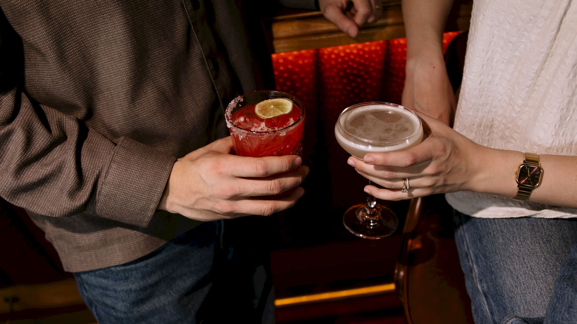 Two people clink cocktails in a dim bar, one with a red drink garnished with a lime, the other a pale cocktail in a coupe.