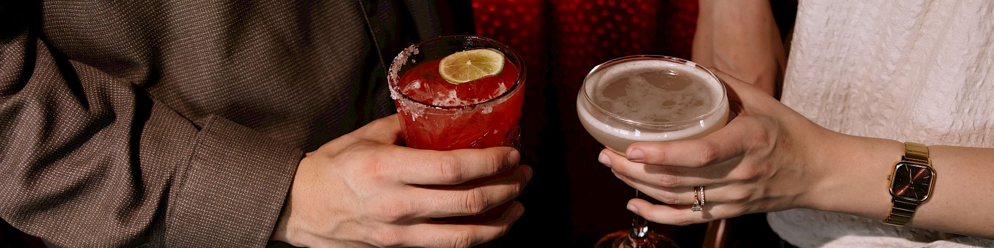 Two people clink cocktails in a dim bar, one with a red drink garnished with a lime, the other a pale cocktail in a coupe.