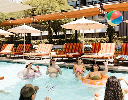 Friends splash in a sunny outdoor pool, sipping drinks by striped loungers under umbrellas and colorful balloons.