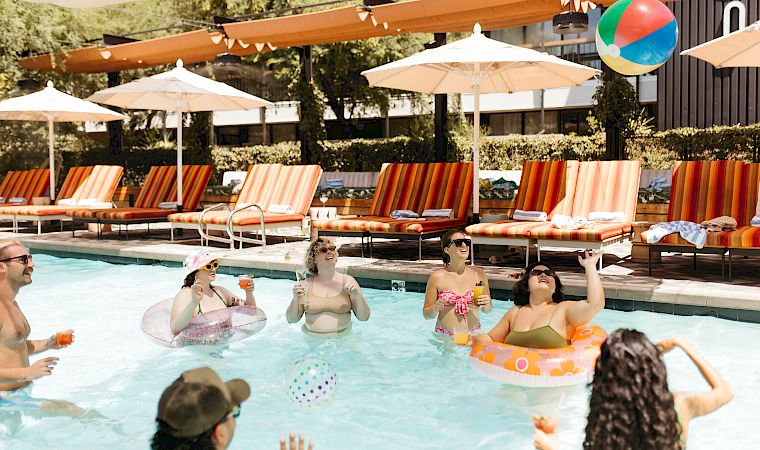 Friends splash in a sunny outdoor pool, sipping drinks by striped loungers under umbrellas and colorful balloons.