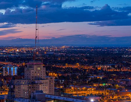 A dusk city skyline with lights, a tall lattice broadcast tower, and stormy clouds over the horizon.