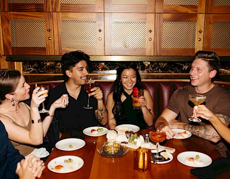 A group of friends in a cozy restaurant booth cheers with beer and laughs over plates at the table.