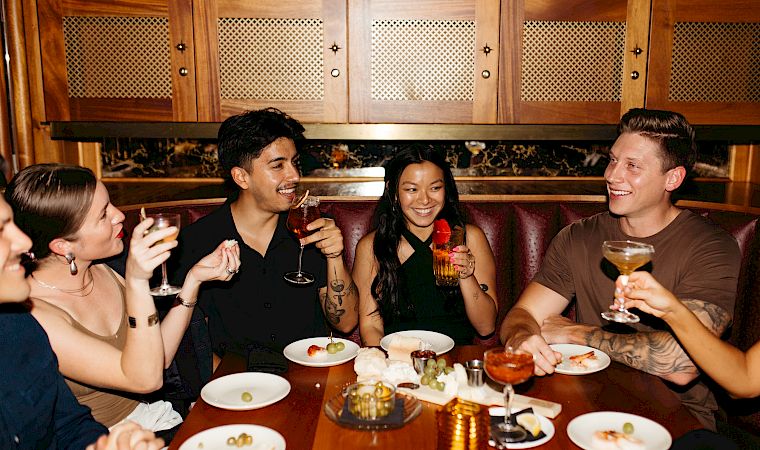 A group of friends in a cozy restaurant booth cheers with beer and laughs over plates at the table.