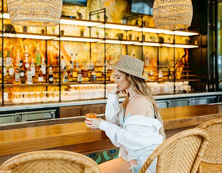 A woman in a straw hat relaxes at a wicker cafe table, sipping a drink under woven pendant lights.