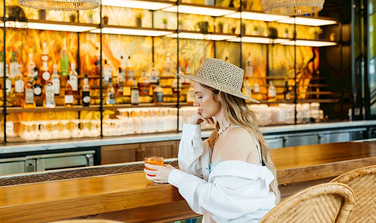 A woman in a straw hat relaxes at a wicker cafe table, sipping a drink under woven pendant lights.