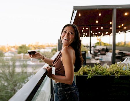 A woman smiles on a patio holding a glass of wine as warm lights glow behind her.