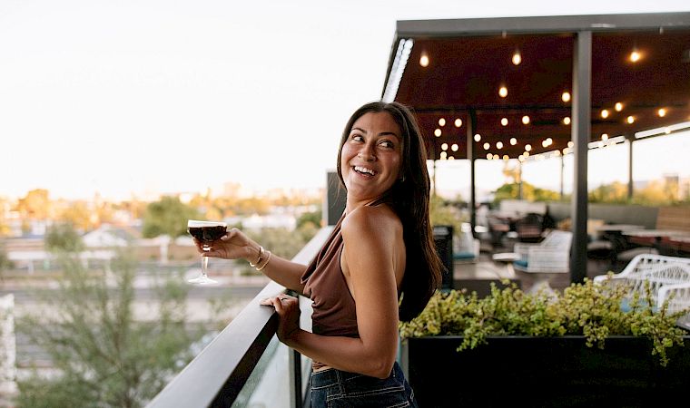 A woman smiles on a patio holding a glass of wine as warm lights glow behind her.