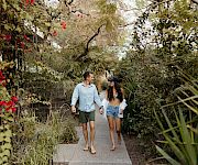 A couple strolls hand in hand along a sunlit garden path, surrounded by trees and red blossoms.