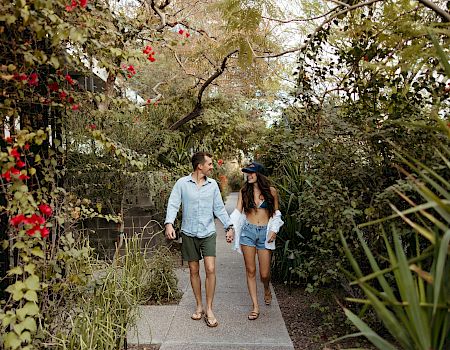 A couple strolls hand in hand along a sunlit garden path, surrounded by trees and red blossoms.