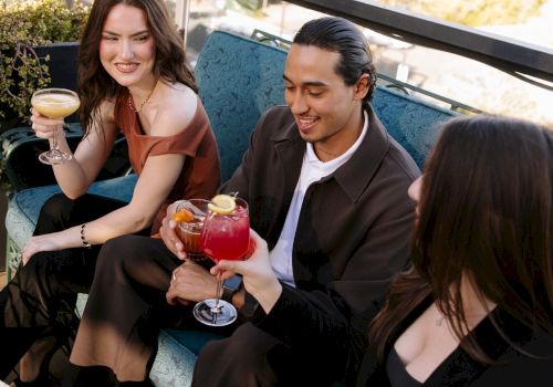 Two friends relax on a patio, smiling and toasting with cocktails at a sunny outdoor table.