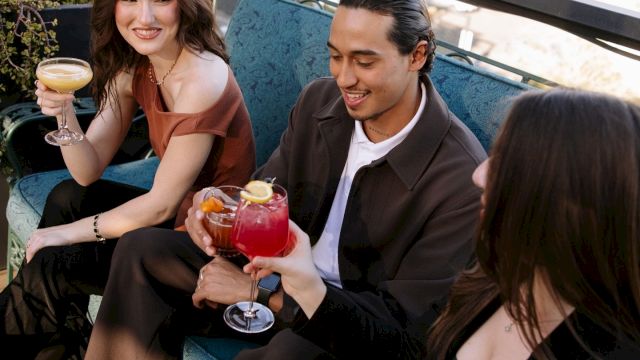 Two friends relax on a patio, smiling and toasting with cocktails at a sunny outdoor table.