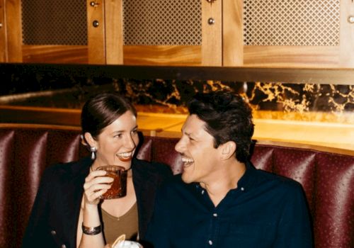 Two people sit in a bar smiling, holding drinks&mdash;one with a beer mug and the other with a glass.