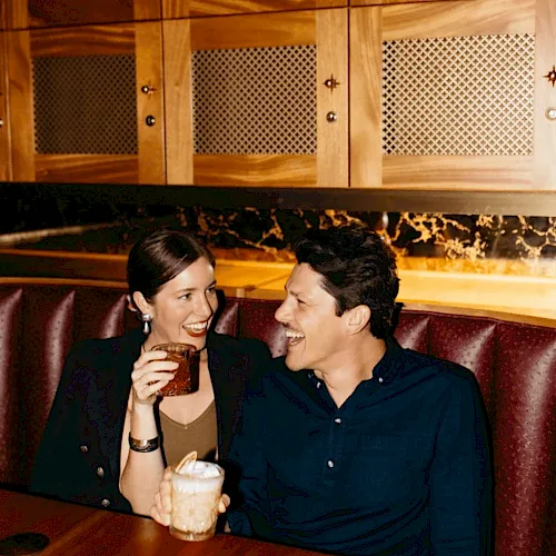 Two people sit in a bar smiling, holding drinks&mdash;one with a beer mug and the other with a glass.