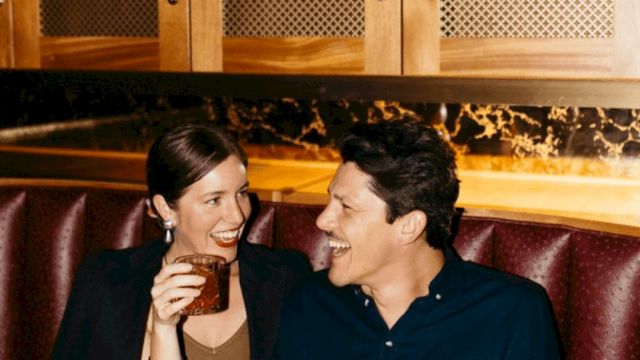 Two people sit in a bar smiling, holding drinks&mdash;one with a beer mug and the other with a glass.