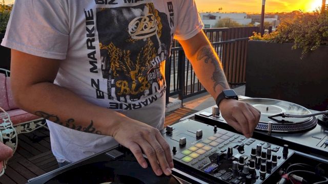 A DJ in headphones spins music on a rooftop deck at sunset, mixing tracks on a turntable/mixer under a golden sky.
