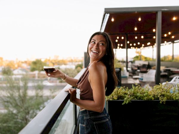 A smiling woman raises a drink on an outdoor terrace at sunset, with warm string lights glowing behind her.