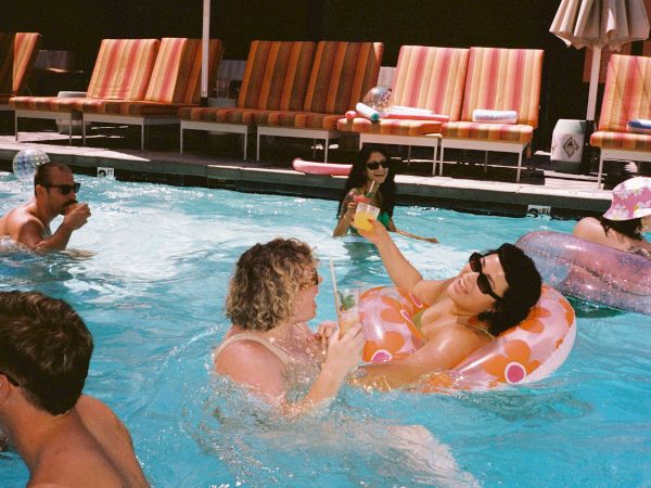 Friends relax in an indoor pool with floaties and drinks, splashing and laughing together.