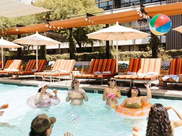 Friends lounging in a sunny outdoor pool, sipping drinks and floating on colorful inflatables at a resort.