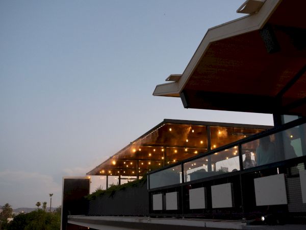 A modern terrace building with warm string lights and a dark red roof, shot at dusk against a pale sky.