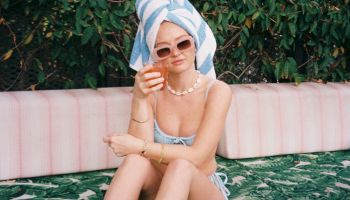 A person with a towel on their head and sunglasses sits on a patterned surface outdoors, with green foliage and a building in the background.