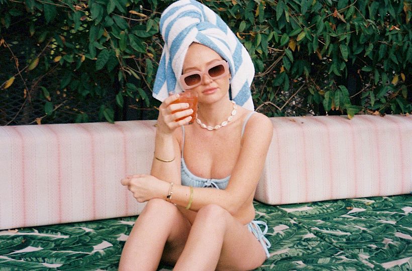 A person with a towel on their head and sunglasses sits on a patterned surface outdoors, with green foliage and a building in the background.