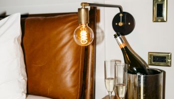 A cozy bedroom setup with a modern lamp, champagne bottle in an ice bucket, two glasses on a wooden nightstand, and citrus-themed wallpaper.