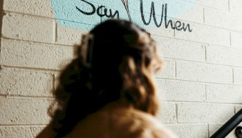 A person facing a white brick wall with a wine glass illustration and the text "Say When" is visible. The person is wearing a light-colored jacket.